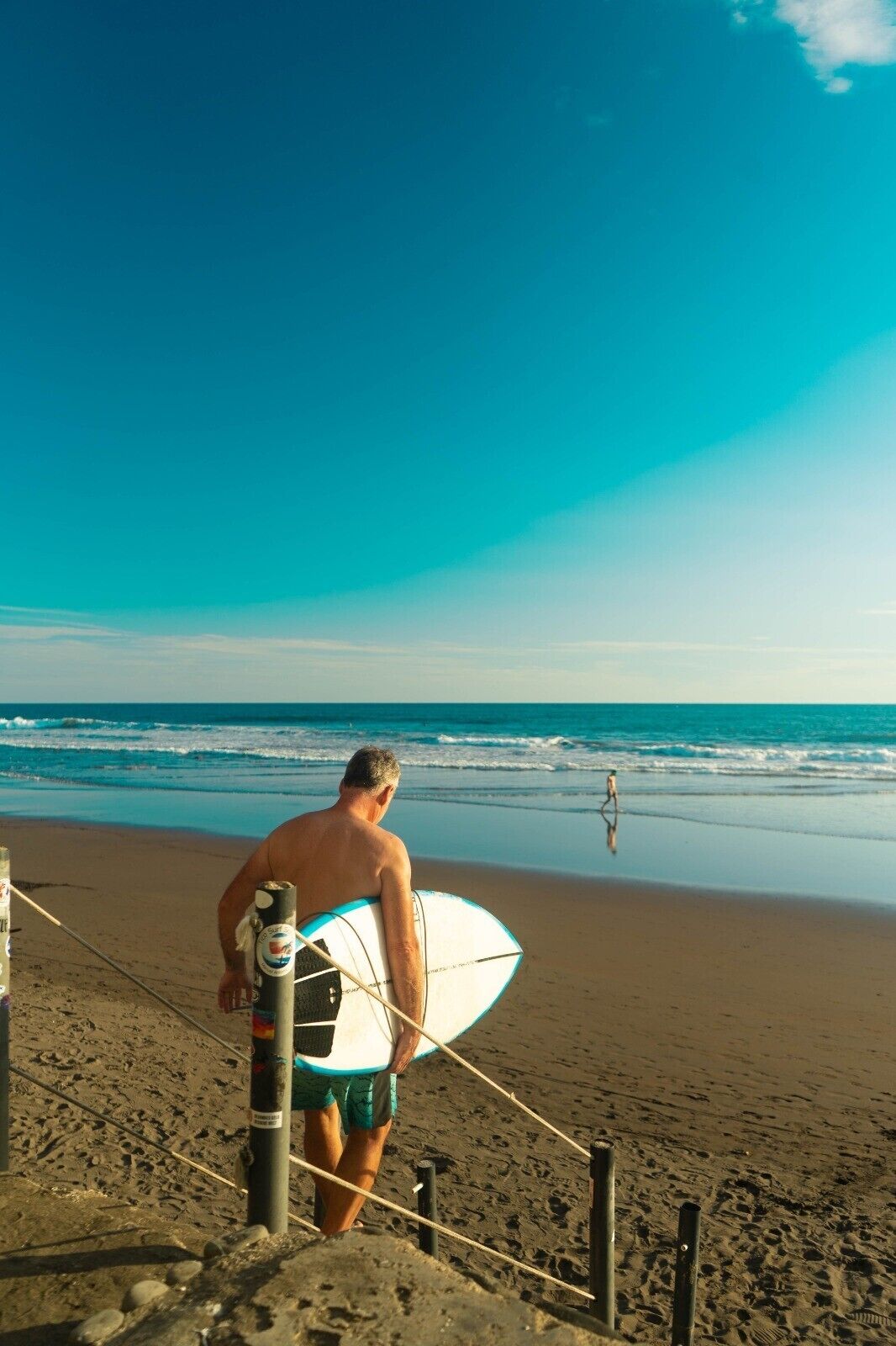 Un hombre lleva una tabla de surf en una playa arenosa, con el océano al fondo.