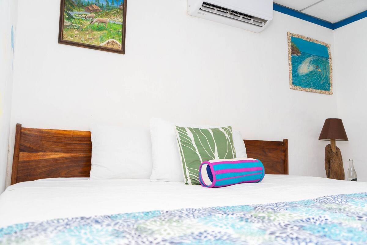 A bed with a patterned bedspread, two pillows, a wooden headboard, an air conditioning unit, and a painting on the wall.