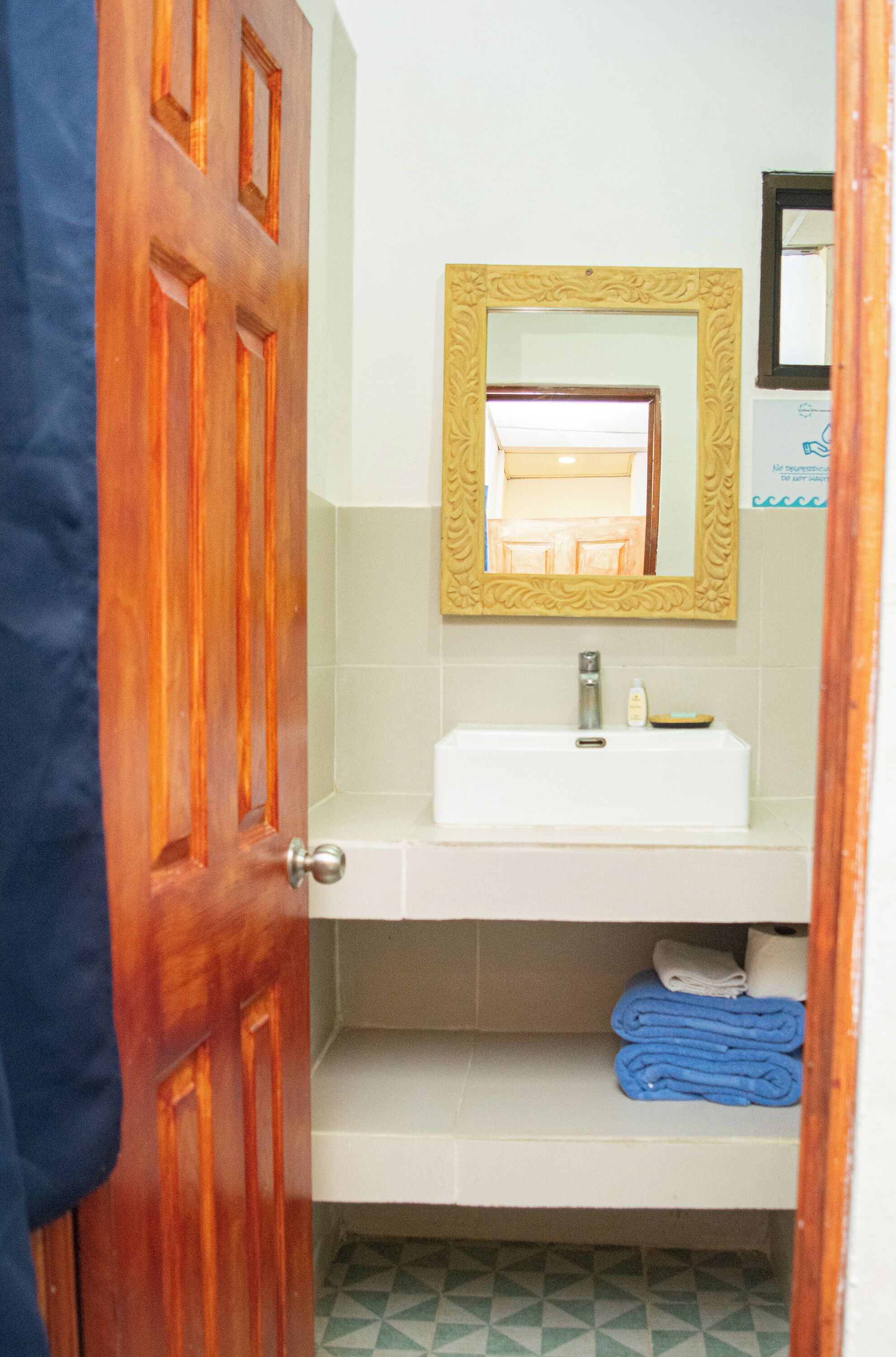 A bathroom with a wooden door, a white sink, a mirror with a decorative frame, and a shelf with folded towels.