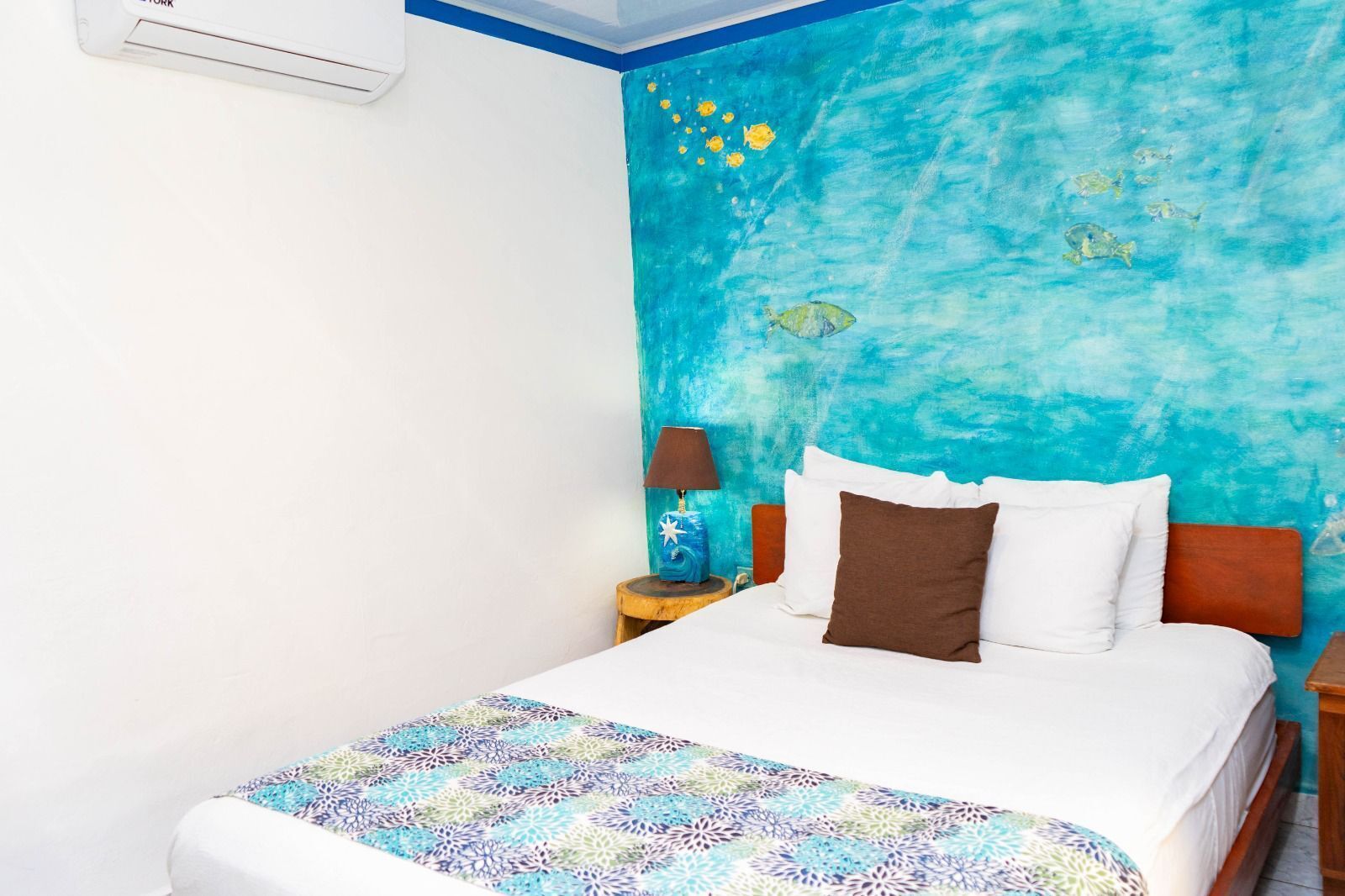 A bedroom with a bed, a nightstand, a lamp, and a wall with a blue and yellow mural.