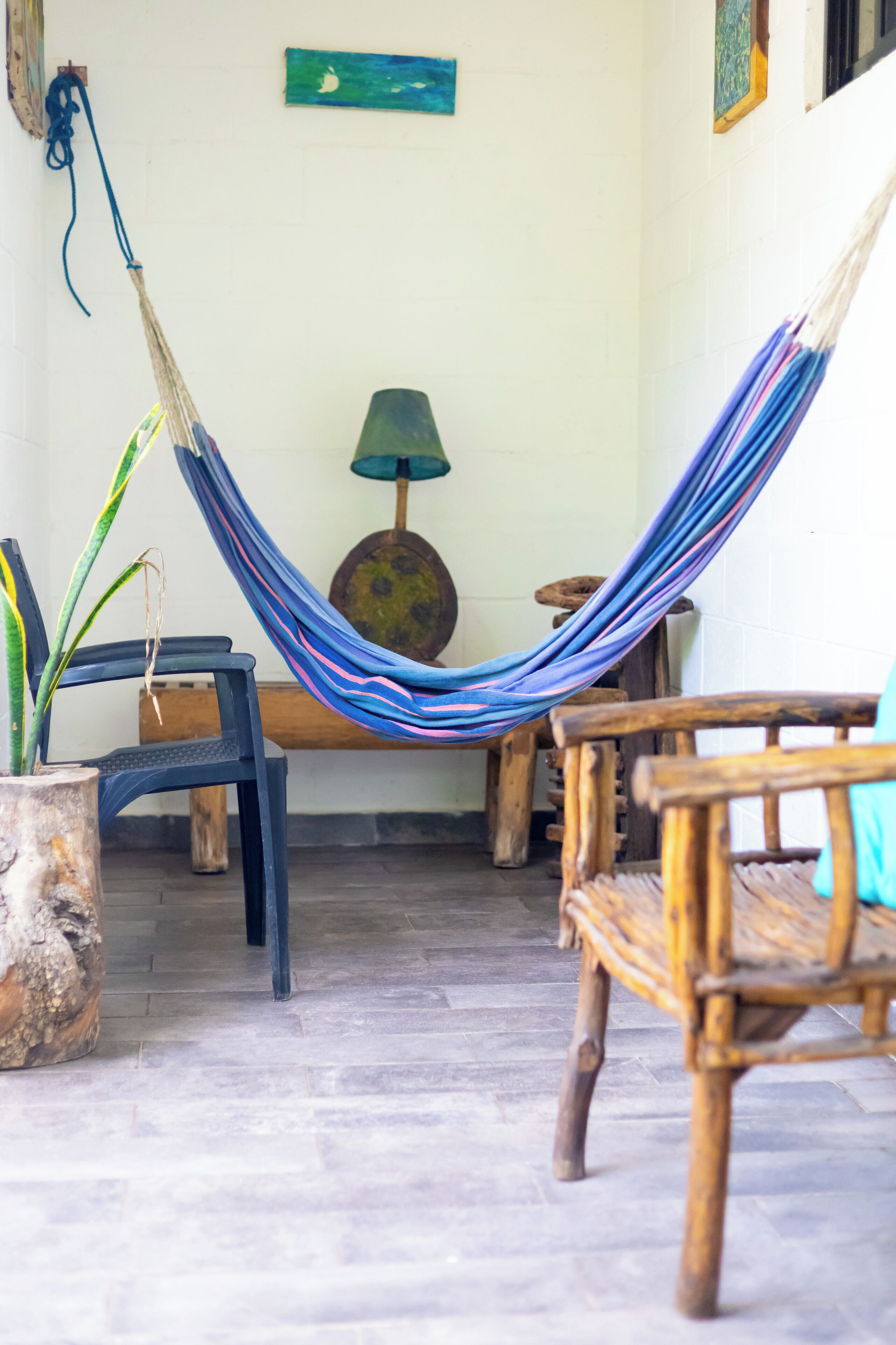 A hammock between two wooden chairs, a potted plant, and a lamp on a tiled floor.