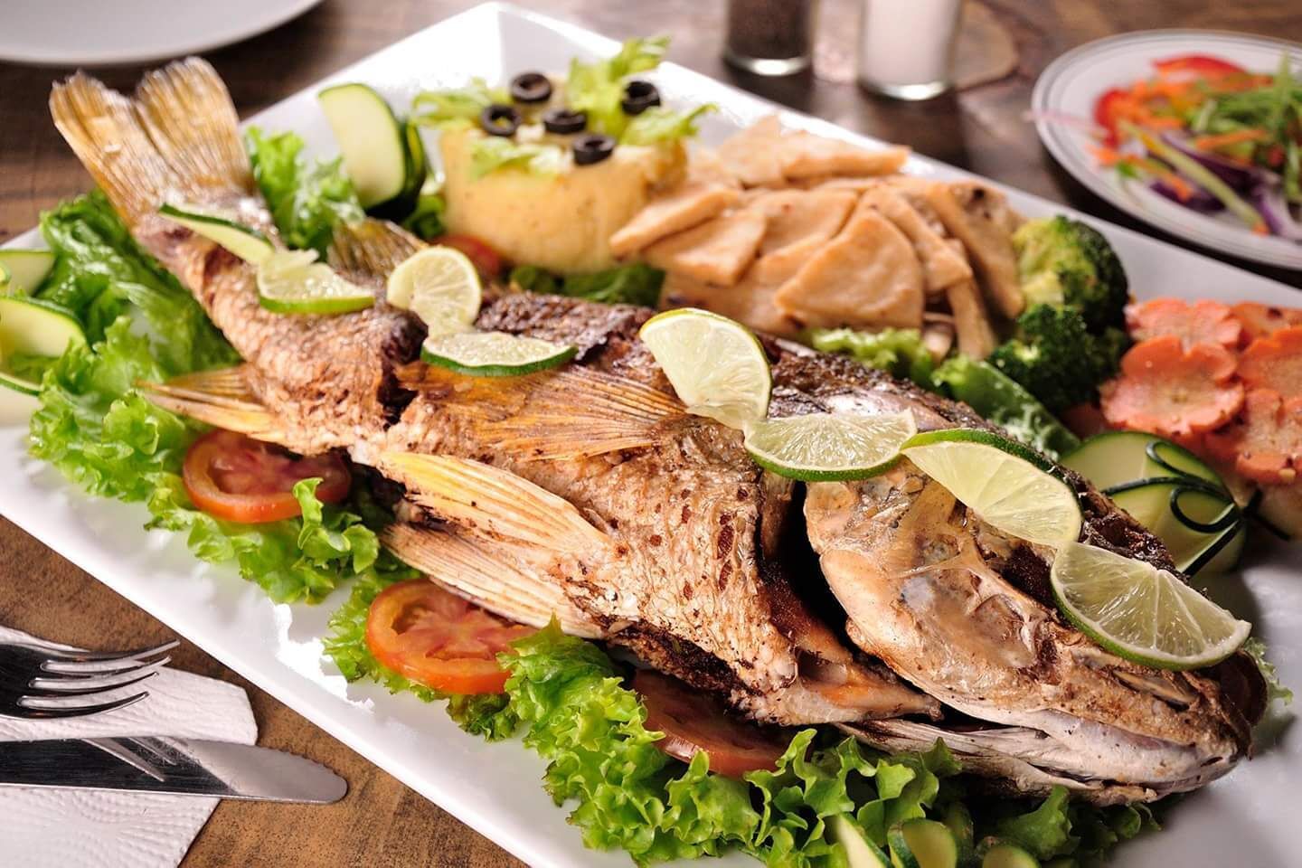 Ein Teller gegrillter Fisch mit Limettenscheiben, Gemüse und Fladenbrot.