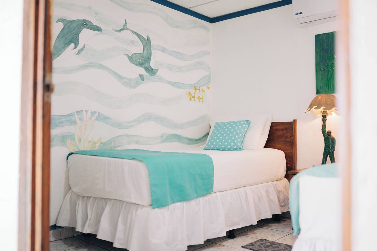 A bedroom with a bed, a nightstand, a lamp, and a wall mural of dolphins.