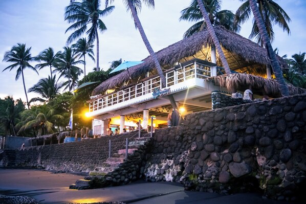 A beachfront hotel with a thatched roof, stone walls, and palm trees.