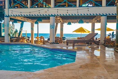 An indoor swimming pool with lounge chairs and umbrellas, surrounded by pillars and a view of the ocean.