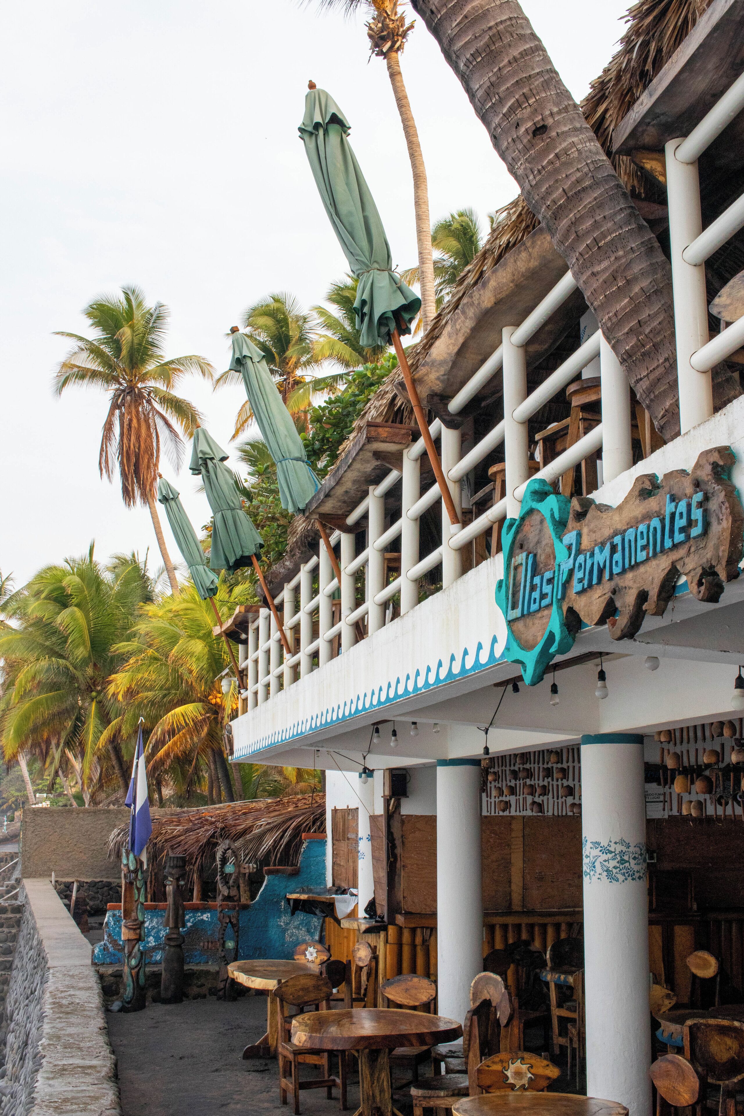 A restaurant with outdoor seating, palm trees, and a sign reading "Las Permanentes".