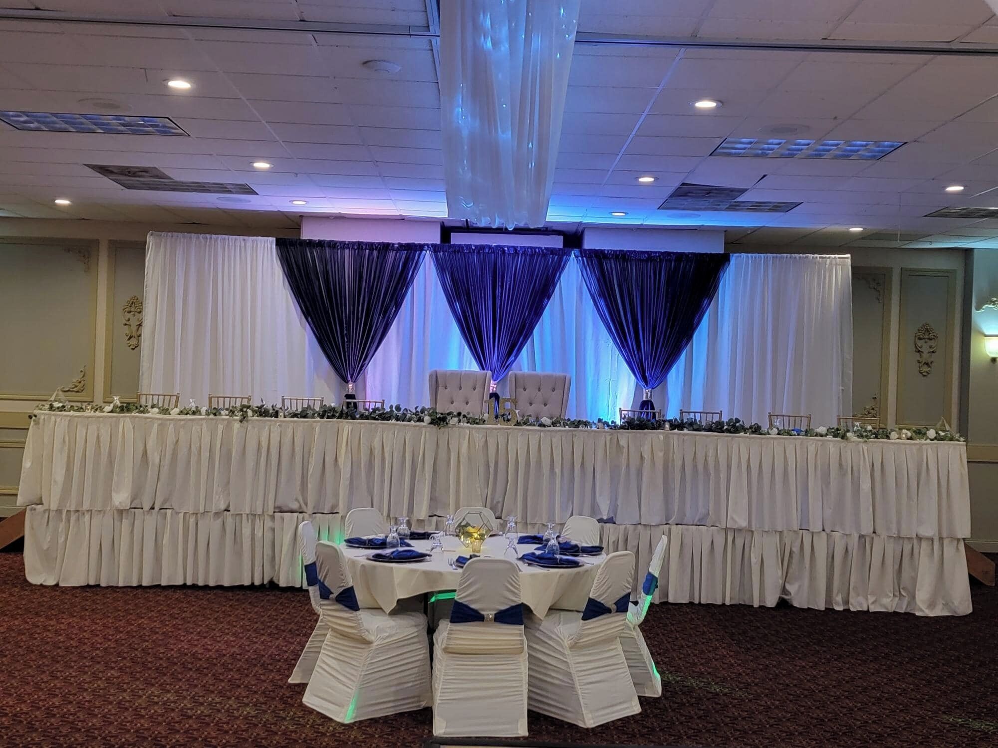 A banquet hall with a long table set for a formal event, draped with white and blue curtains, and a carpeted floor.