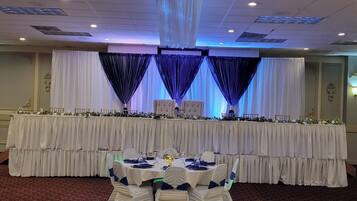 A banquet hall with a long table set for a formal event, draped with white and blue curtains, and a carpeted floor.