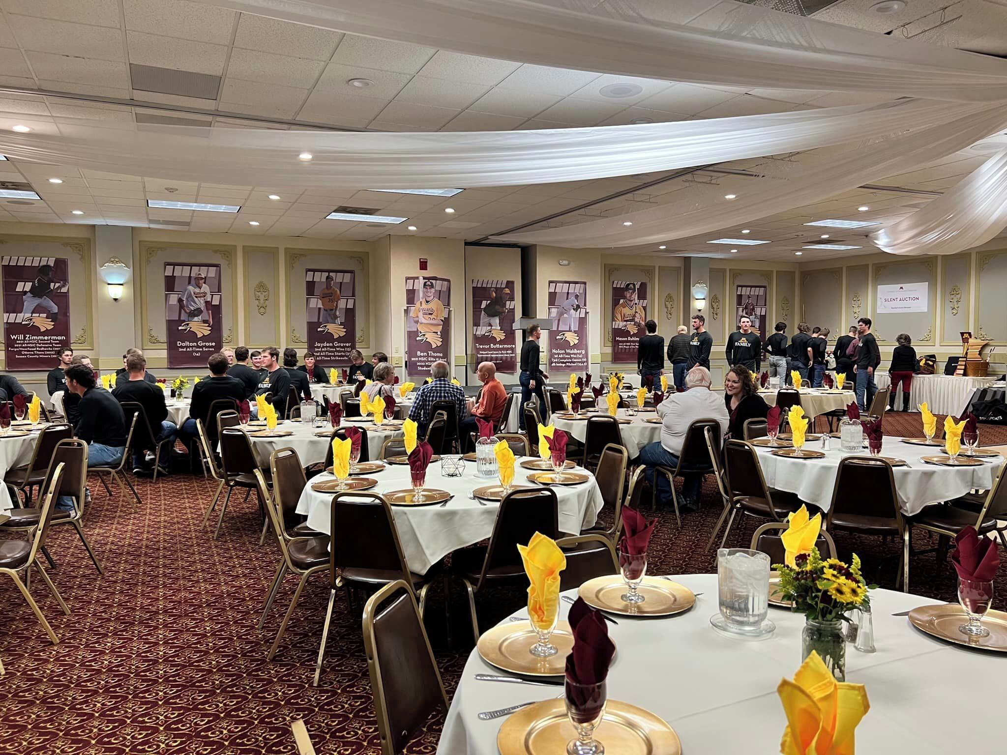 Een feestzaal met ronde tafels gedekt voor een diner, met gele en bordeauxrode servetten, en een vloerbedekking.