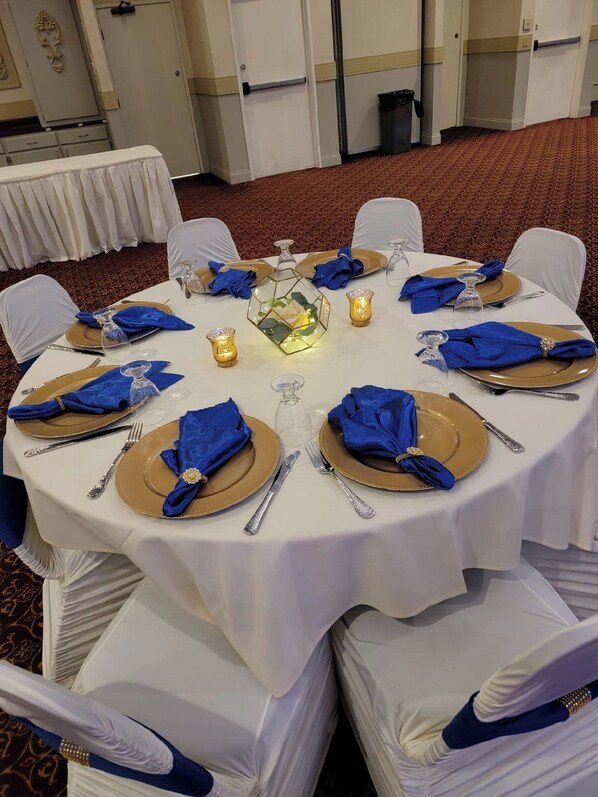 Une table ronde dressée pour un événement formel, avec des nappes blanches, de la vaisselle dorée et bleue, et des bougies.