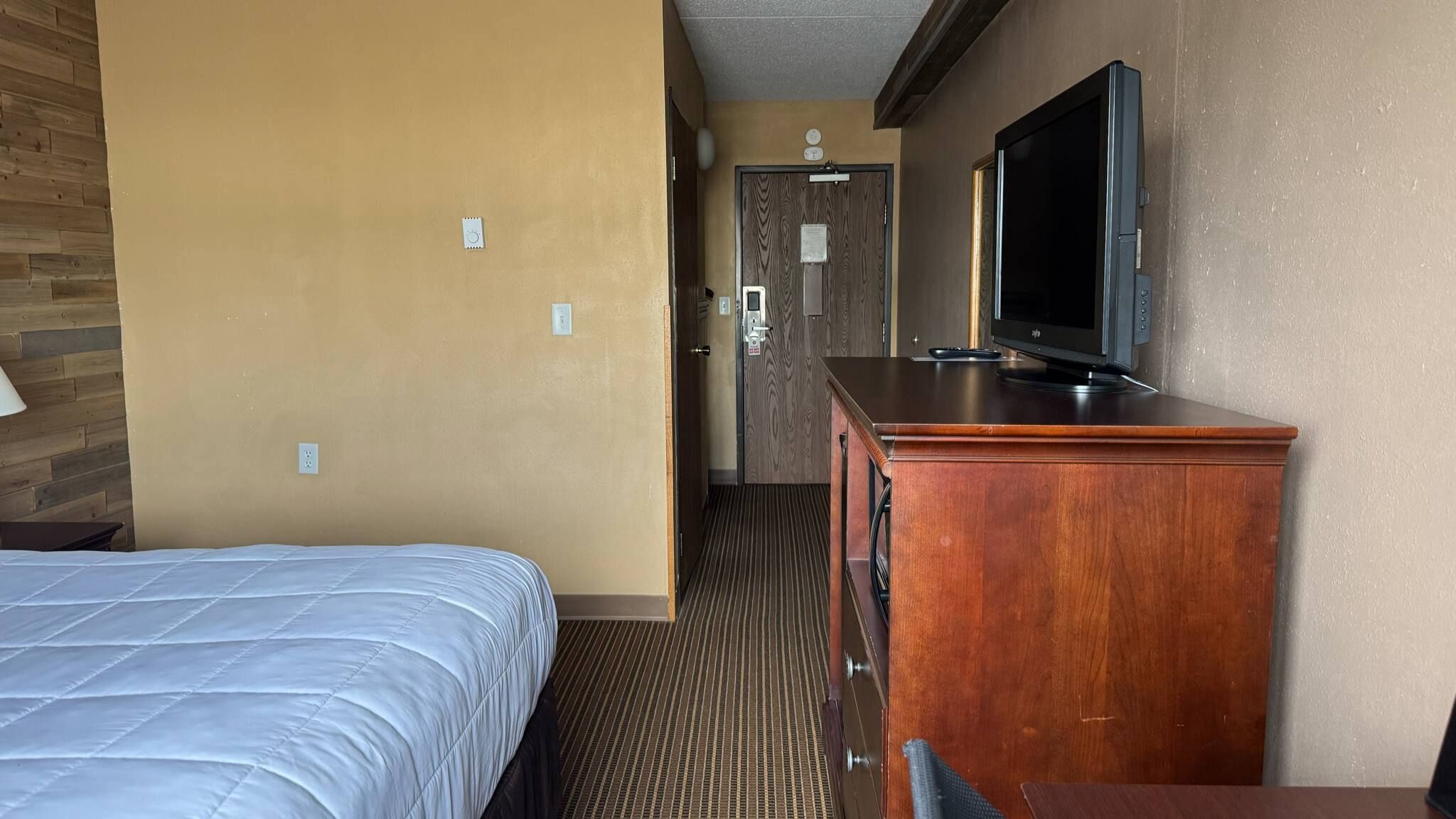 A hotel room with a bed, a wooden dresser, a television, and a door leading to a hallway.