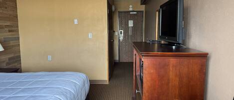 A hotel room with a bed, a wooden dresser, a television, and a door leading to a hallway.