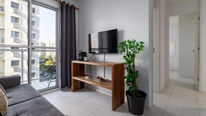 A modern living room with a large window, a flat-screen TV, a wooden coffee table, a grey sofa, and a potted plant. - Charm in Barra da Tijuca Great Wifi Sa508 (Rio de Janeiro)