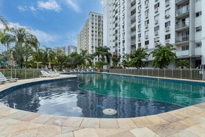 Ein Swimmingpool mit Liegestühlen und einer gefliesten Terrasse, umgeben von hohen Wohnhäusern und Palmen.