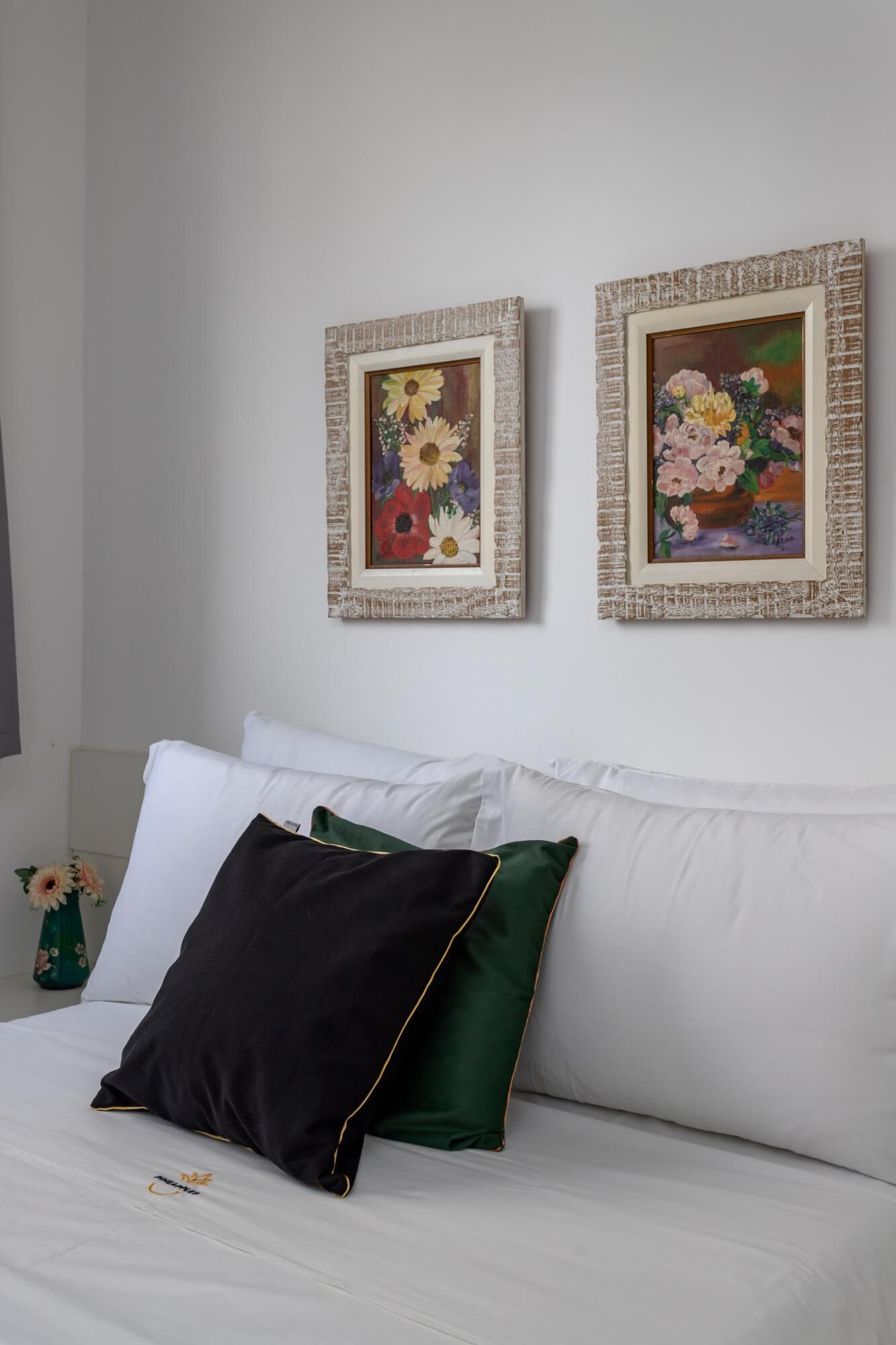Un dormitorio con una cama, almohadillas blancas y dos cuadros enmarcados con motivos florales en la pared.