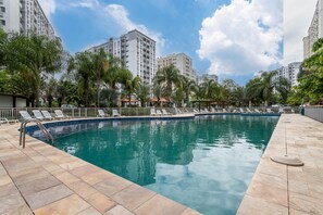 Ein Swimmingpool mit Liegestühlen, umgeben von Palmen und hohen Gebäuden.