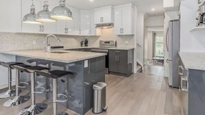 Una cocina moderna con armarios blancos, una isla central con taburetes, electrodomésticos de acero inoxidable e iluminación colgante.