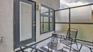 Un área de estar al aire libre con paredes de vidrio, sillas y mesa de metal, una puerta con panel de vidrio satinado y una ventana con vistas a un edificio.
