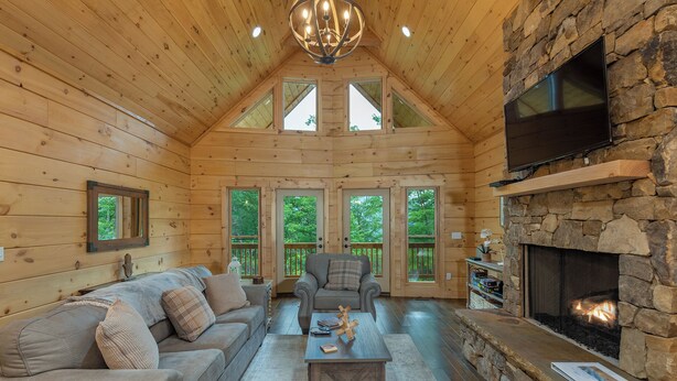 Ein Blockhaus-Interieur mit Kamin aus Stein, einem an der Wand befestigten Fernseher und einer gemütlichen Sitzecke mit Sofa und Sesseln.