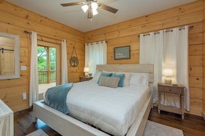 A wooden-paneled bedroom with a bed, nightstands, a ceiling fan, and a view of the outdoors. - Dream Mtn~Hot Tub+Fire Pit+Grill~Fireplace & WiFi! (Black Mountain)