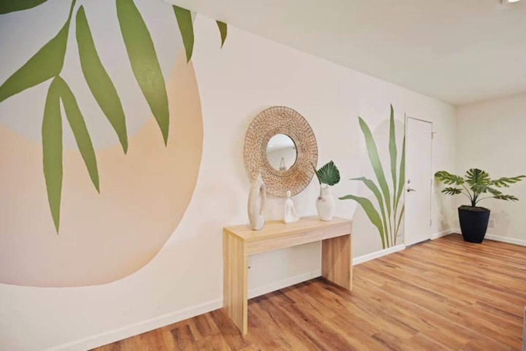 A room with a wooden floor, a round mirror on a wooden console, and a large wall mural of green leaves.