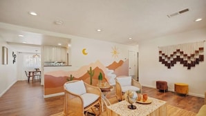 Een woonkamer met een houten vloer, een salontafel, twee rieten fauteuils, een vloerkleed en een wanddecoratie met een woestijnlandschap.