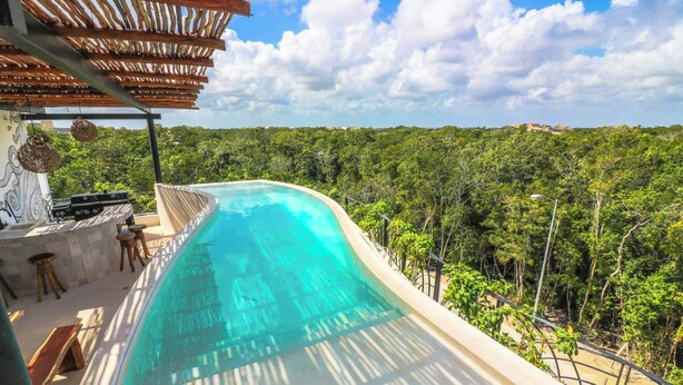 En buet infinity-pool med udsigt til tæt skov, grillområde og pergola.