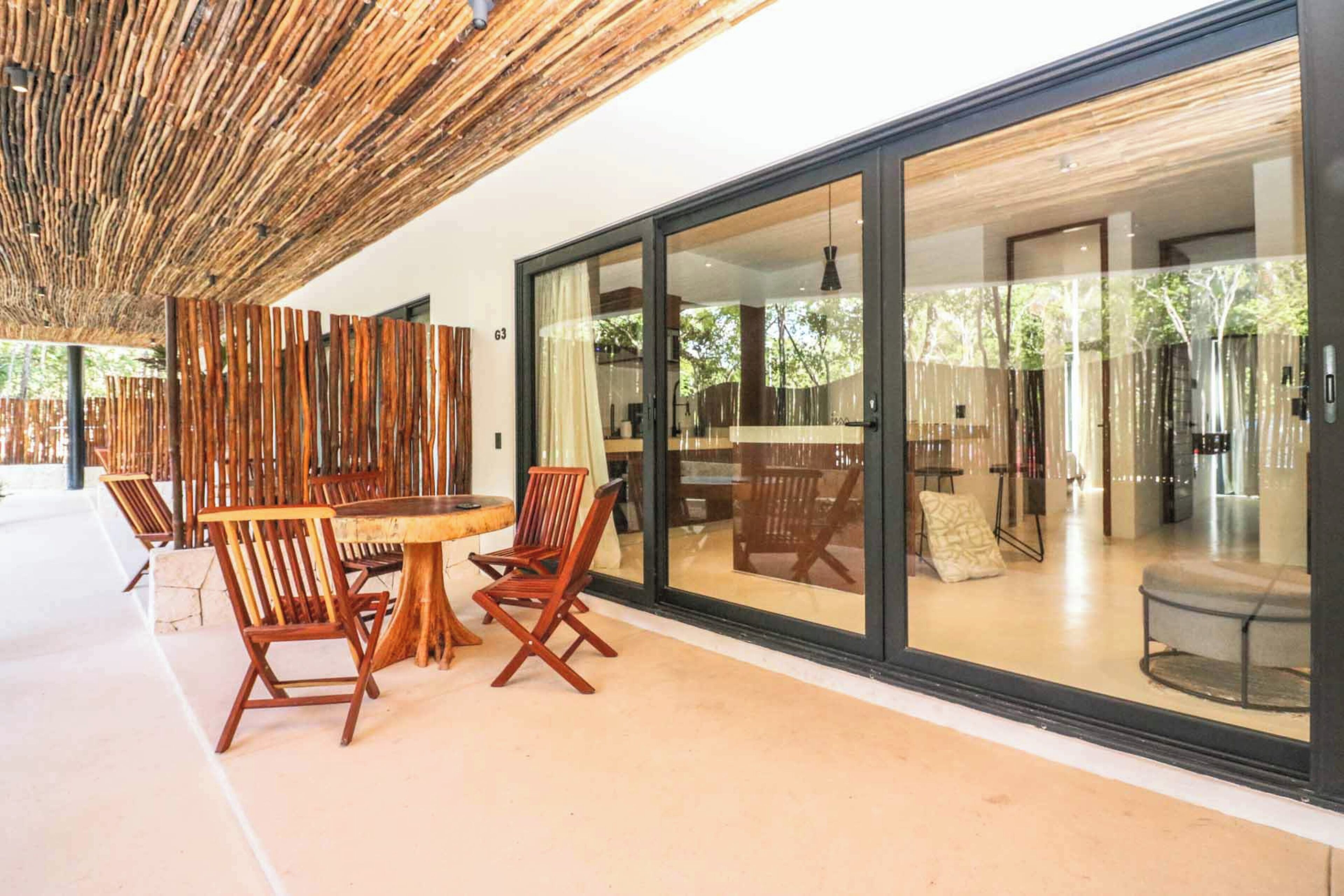 Un área de comedor al aire libre con mobiliario de madera y una pared de vidrio que da a un interior moderno.
