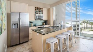 Una cocina moderna con una isla grande, electrodomésticos de acero inoxidable y vistas a palmeras a través de puertas correderas de vidrio.