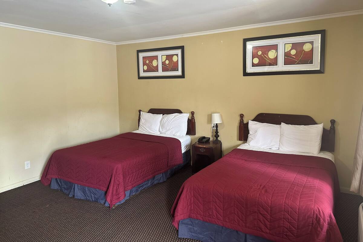 A hotel room with two beds, each with a red bedspread and white pillows. There are framed pictures on the wall, a bedside table with a lamp, and a ceiling light.