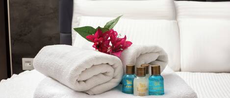 A neatly folded stack of white towels on a textured white bedspread, accompanied by two bottles of Lenus hotel shampoo and conditioner.