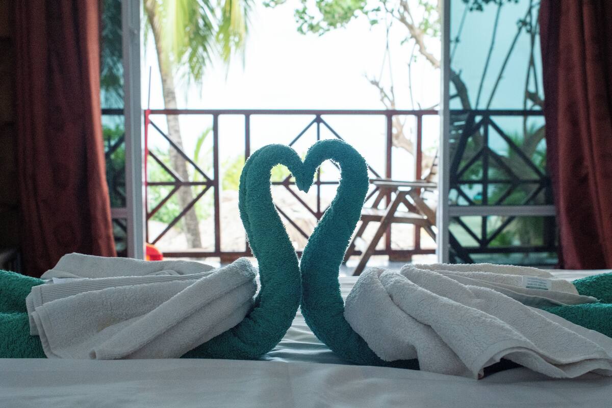 A bed with towels arranged in a heart shape, a window with a view of palm trees, and a balcony with a railing.