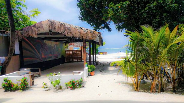 Una estructura frente al mar con techo de paja, rodeada de plantas en macetas y palmeras, que da a una playa arenosa.