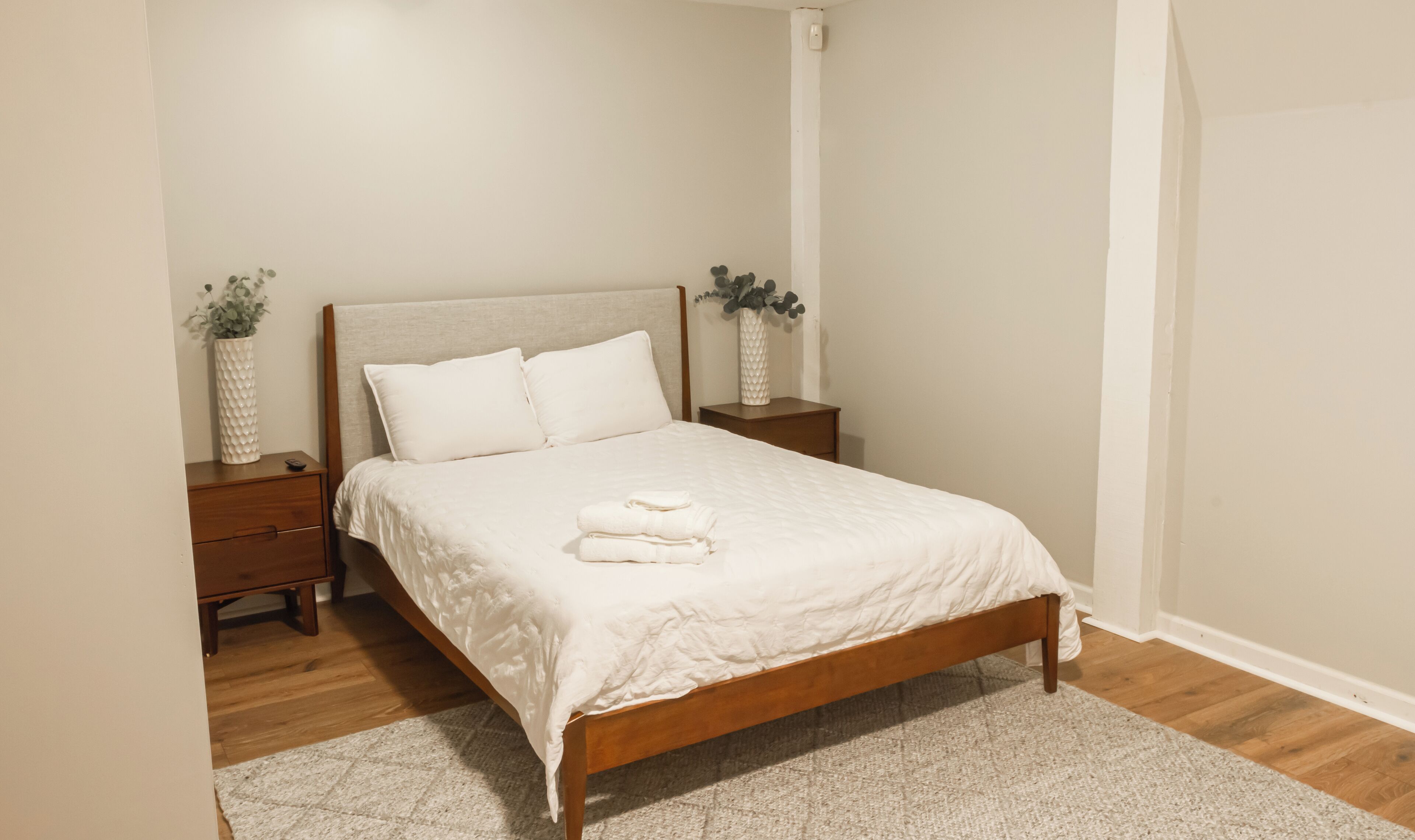 A neatly made bed with white bedding, a wooden headboard, and a small bedside table with a vase of flowers.