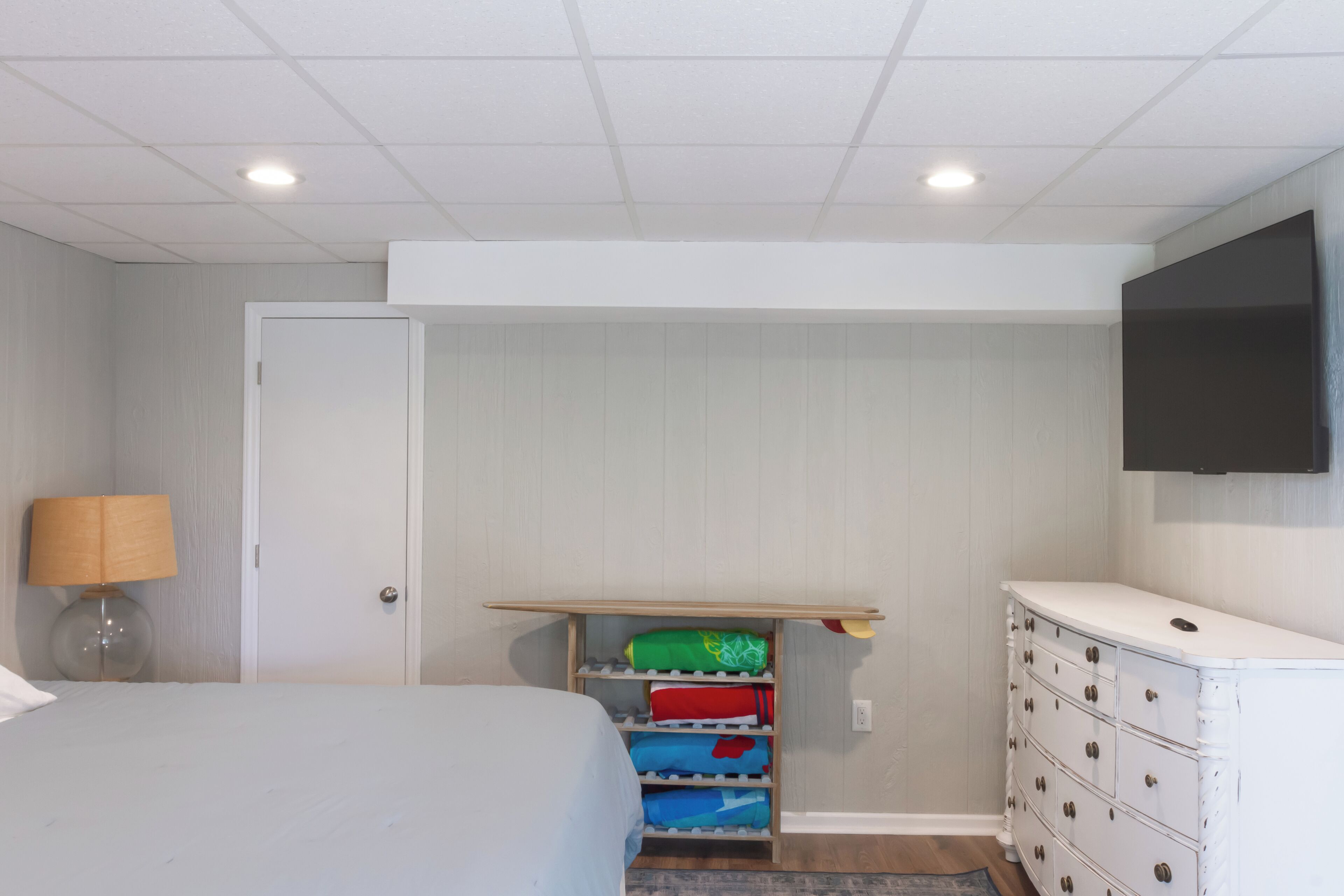 Un dormitorio con cama, cómoda, televisor montado en la pared y un mueble de almacenamiento con contenedores de colores.