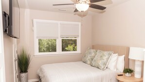 Quarto com cama, ventilador de teto, janela com persianas, planta em vaso e mesa de cabeceira com abajur.