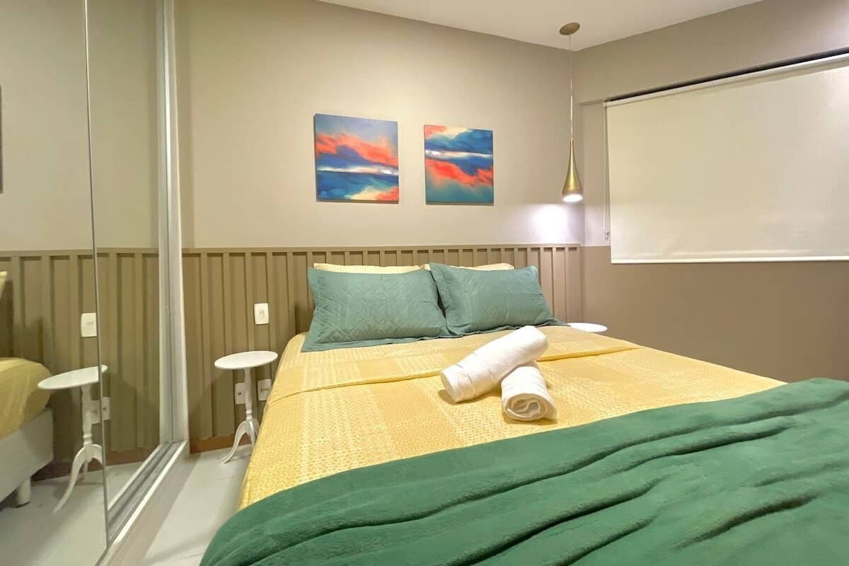 A hotel room with a bed, two pillows, a rolled towel, a bedside table, a mirror, and two paintings on the wall.