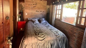 Un dormitorio con techo de madera, una cama con colcha estampada, una mesita de noche roja, una pared con un mensaje y una ventana con vistas.