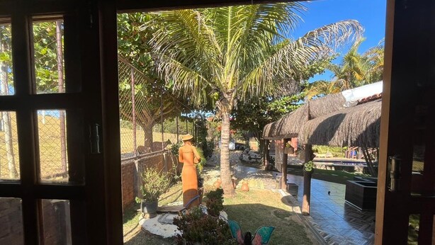 Vista a través de una ventana de un jardín tropical con una estructura de techo de paja, una palmera y una estatua.