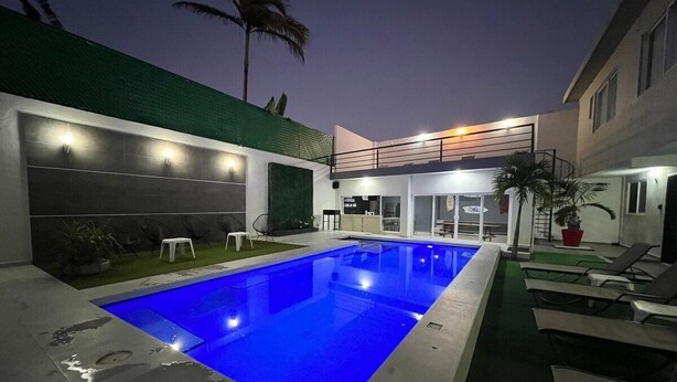 Una piscina moderna al aire libre con una fuente iluminada en azul, rodeada de sillones de descanso y una terraza bien iluminada.