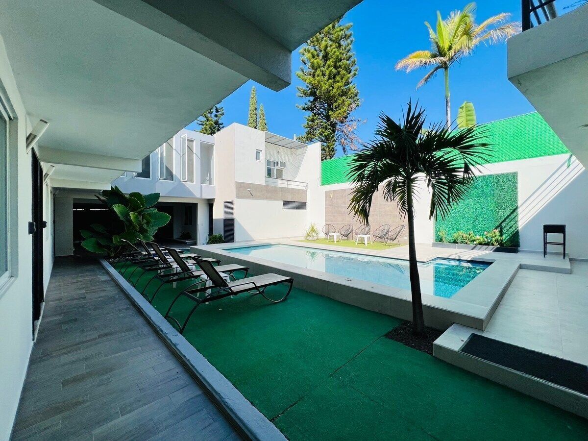 A modern outdoor pool area with a green artificial turf, lounge chairs, and a palm tree.