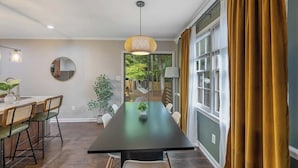 A modern dining area with a long table, chairs, a hanging light fixture, and a view of the outdoors. - Outdoorsy ~ Fresh Spring Vibes ~ Sauna & Cold Plunge ~ 10min Downtown (Raleigh)