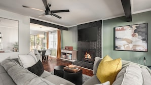 A modern living room with a fireplace, a ceiling fan, and a dining area visible in the background. - Outdoorsy ~ Fresh Spring Vibes ~ Sauna & Cold Plunge ~ 10min Downtown (Raleigh)