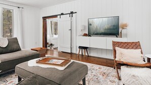 A modern living room with a sofa, ottoman, and a television on a wall. - Muskoka Zen, Outdoor Jacuzzi Spa, Ski or Hike 3 Bedroom 2 Bath (Huntsville)