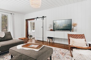 A modern living room with a sofa, ottoman, and a television on a wall. - Muskoka Zen, Outdoor Jacuzzi Spa, Ski or Hike 3 Bedroom 2 Bath (Huntsville)