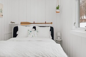 A bed with a white quilt, two pillows with a leaf pattern, a wooden paddle on the headboard, a small potted plant, and a bedside table with a lamp. - Muskoka Zen, Outdoor Jacuzzi Spa, Ski or Hike 3 Bedroom 2 Bath (Huntsville)