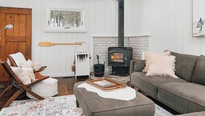 A cozy living room with a fireplace, a wooden door, a window with a view of trees, and a couch with cushions. - Muskoka Zen, Outdoor Jacuzzi Spa, Ski or Hike 3 Bedroom 2 Bath (Huntsville)