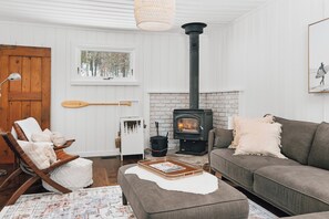 A cozy living room with a fireplace, a wooden door, a window with a view of trees, and a couch with cushions. - Muskoka Zen, Outdoor Jacuzzi Spa, Ski or Hike 3 Bedroom 2 Bath (Huntsville)