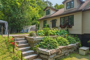 A house with a stone staircase leading to a porch, surrounded by lush greenery and a hammock. - Muskoka Zen, Outdoor Jacuzzi Spa, Ski or Hike 3 Bedroom 2 Bath (Huntsville)