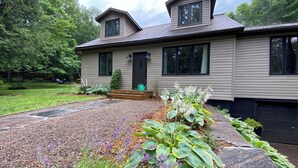 A two-story house with a front porch, a small garden with various plants, and a gravel driveway. - Muskoka Zen, Outdoor Jacuzzi Spa, Ski or Hike 3 Bedroom 2 Bath (Huntsville)
